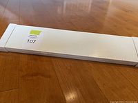 Long white rectangular box laid flat on wooden floor, shows unopened IKEA Mosslanda shelving package with seller lot sticker.