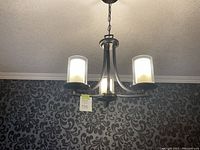 Black brown metal 3-light chandelier with frosted cylindrical glass shades, hanging from ceiling on chain, against decorative wallpaper.