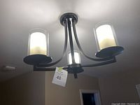 Front and side view of the black brown three-arm ceiling light fixture with frosted glass cylindrical shades illuminated.