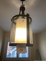 Pendant light installed and turned on, showing warm light through frosted glass cylinder inside black brown metal frame with clear glass panels.