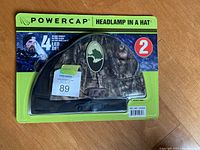 Front packaging view showing camouflage hat with built-in headlamp and branding.