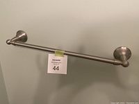 Brushed nickel towel bar mounted on a wall with its mounting brackets visible on both ends and a lot number tag attached to the bar.