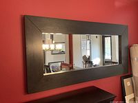Full length rectangular mirror mounted on a red wall above a dark wood cabinet. Shows the dark brown wide beveled solid wood frame and the reflection of the room.