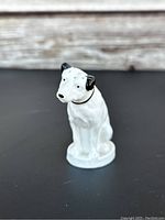 White ceramic Nipper dog mascot figure with black ears, seated on a round base, shown against wood background