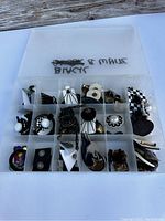 A photo showing a clear plastic divided box containing black and white themed earrings in various styles such as studs, dangles, and clip-ons, some with pearl-like beads and floral motifs.