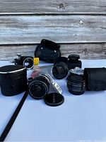Photo showing multiple Miranda camera lenses, Leica lens cap, two black leather cases, and small accessories on wooden surface.