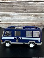 Side view of blue police van showing police logo, barred windows on rear, white wheels, and partial missing parts.
