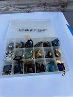 Top view of open plastic organizer box showing 18 compartments filled with various purple and teal earrings of different styles and shapes.