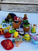 Overview of vintage McDonald’s toys including Mr. Potato Head figures, frog figurines, plastic vehicles, firefighter hat, and other small toy accessories arranged on a white background.
