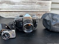 Front view of Minolta SR3 camera with attached lens, vintage light meter to the left and black leather carrying case to the right on a black surface in front of wooden background.