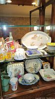 Glass cabinet shelf showing ceramics, glasses, and cups including Lipton advertising glasses and various decorative ceramic plates and bowls.