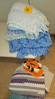 Stack of three crocheted lap blankets on a white piece of furniture. Top blanket is cream with pastel colors, middle one is blue and purple with fringes, bottom is not fully visible.