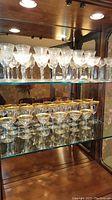 Display cabinet shelf showing two rows of etched floral pattern wine glasses on upper glass shelf and glasses with gold rim on lower shelf.