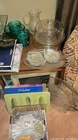 Photo showing green glass bowls, clear glass pitcher, clear glass cake stand with dome, clear textured glass platter, cream-colored dishes, and assorted glassware on small table next to couch and carpeted floor.
