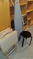 Side view of the folding ironing board with blue striped cover, two light wood folding TV tables, small white folding metal table, and black stool
