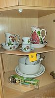 Shelf displaying floral porcelain salad bowl and matching pitcher, tea cups with saucers, and white mixing bowls below with utensils.