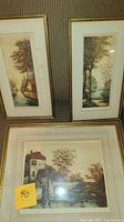 Three framed watercolor paintings with landscape scenes: house by river, tall trees by lake, and stone bridge over water.