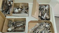 Four cardboard boxes filled with assorted silverware including forks, spoons, and knives.
