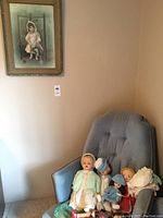 Full view of the framed picture and the grouping of vintage dolls on a grey upholstered chair against a beige wall.