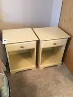 Pair of cream nightstands placed side by side on carpeted flooring against wall, showing overall condition and design.