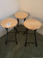 Three light wood round top folding stools arranged together showing the wood seat surfaces and black folding metal frames.