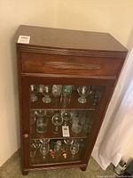 Front view of vintage wooden china cabinet showing top drawer, glass door, and three glass shelves with glassware inside.