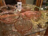 Top view of various pink depression glass bowls with distinct decorative rims and yellow etched dessert cups on glass shelf.