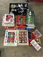 Overview of entire lot showing storage box with bubble-wrapped items, multiple boxed ornament sets, and green box of string lights