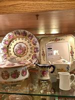 Shelf with ornate large plate featuring a romantic scene, square floral tray, floral tureen, and two creamers including one white ribbed