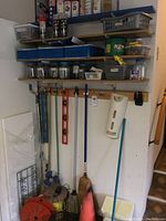 Overview photo showing wall shelves with labeled boxes and jars containing small hardware items, spray paint cans on top shelf, hanging cleaning tools and levels, a broom and reel of red rope on the floor.