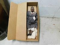 Full view of the doll laying inside the brown cardboard box showing clothing and packaging.