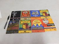 Top view of the 10 vintage large matchbooks arranged in two rows with a pen included for scale. Matches inside are unstruck and matchbooks are clean and in good vintage condition.