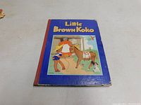 Front hardcover of the Little Brown Koko book showing illustrated cover with child, horse, and dog in color against blue background with red spine.