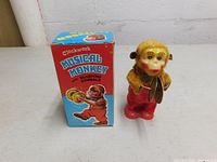 Front view of the vintage musical monkey toy holding cymbals next to the original box showing colorful graphics and text.