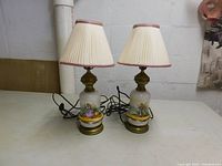 Two antique porcelain lamps with pleated shades, showing the pair together from the front with electrical cords.
