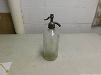 Full view of vintage glass seltzer bottle with metal siphon top on table.