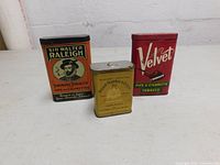 Three vintage tobacco tins arranged in a row showing front labels: Sir Walter Raleigh, Philip Morris & Co., Velvet.
