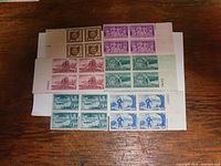 Photo shows six blocks of four uncancelled U.S. commemorative stamps arranged on a wooden surface. Stamp subjects include Ohio Sesquicentennial, American Bar Association, Lewis and Clark Expedition, Washington Territory, Future Farmers of America, and Centennial of Opening of Japan.