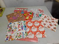Flat lay of multiple colorful vintage Christmas wrapping paper sheets spread out showing a variety of festive designs including Santa Claus, birds, children, and holly.
