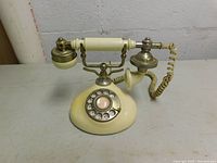 Front facing photo of vintage rotary dial phone in cream and brass, showing dial, handset, coiled cord.