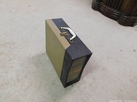 Full view of the vintage portable record player showing its rectangular shape, olive green and black exterior, and white carrying handle on top.