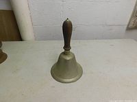 Photo of the vintage school bell standing upright on a white surface, showing the full bell and wooden handle.