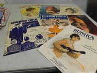 Photo of multiple vintage sheet music pieces laid out on a table, visible titles including 'HUNIKA LULLABY', 'CHICKERY CHICK', 'EGYPTIAN NIGHTS'