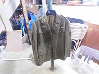 Full view of the olive drab WWII U.S. Army jacket hanging on a stand, showing front with pocket flaps and buttons