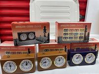 Five analog chess clocks in assorted colors with their original boxes, showing front faces and packaging for identification.