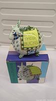 Front and side view of the goat-shaped ceramic coin bank with colorful patchwork design and text on yellow banner.