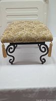 Top front angle view of the foot stool showing patterned cushion with tassels and metal legs.