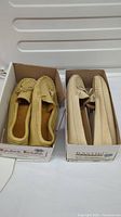 Two pairs of leather moccasins in their original boxes. The pair on the left is tan colored with lace, the pair on the right is a lighter beige with a smooth finish.