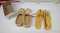 Photo showing both pairs of moccasins side by side, with one pair in a box and the other displayed next to it, highlighting difference in color and material.