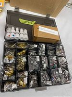Top view of a transparent compartmentalized box showing multiple small bags of jewellery craft accessories in gold and silver tones, along with bottles and a box of water slide decal paper.